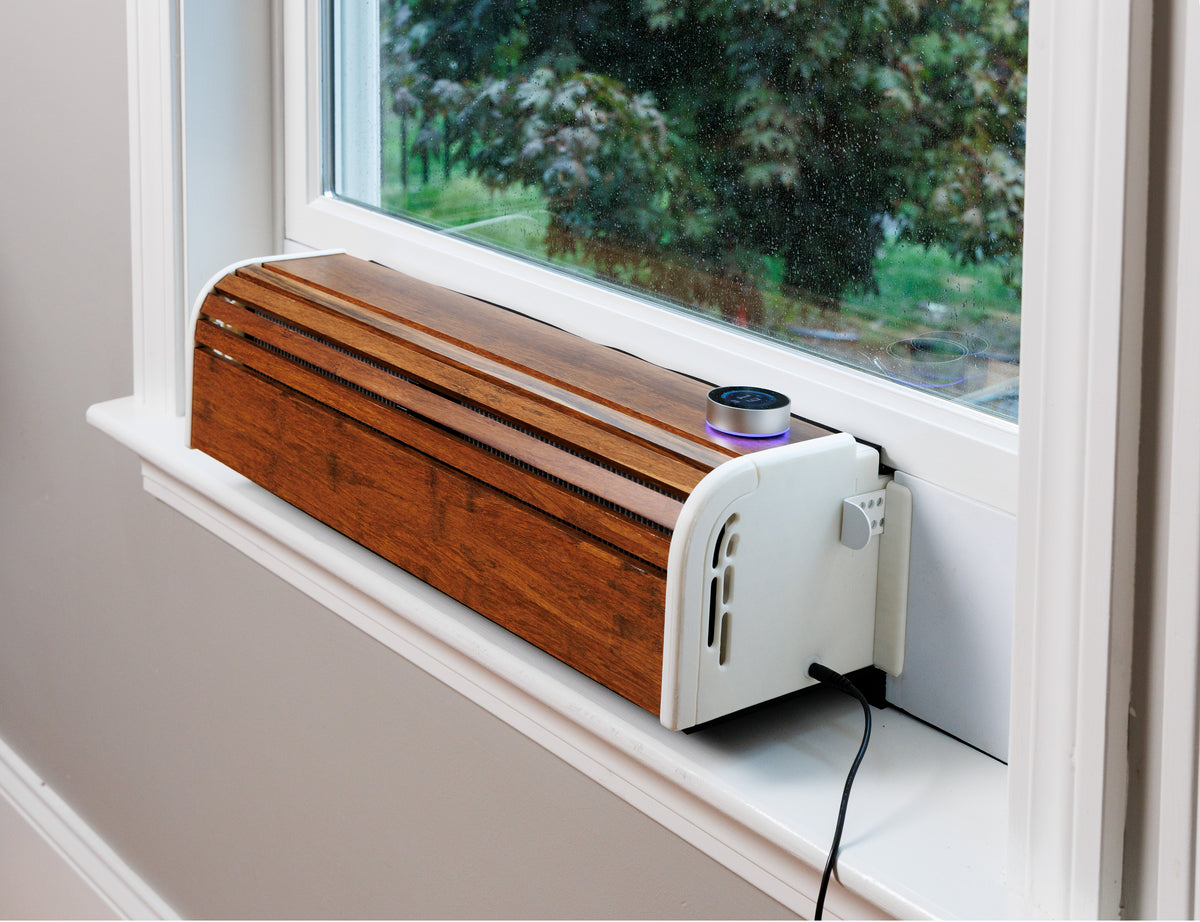 A wood-look window ERV air exchanger with a white side panel and a black power cord, placed on a white windowsill next to a window showing green trees outside.