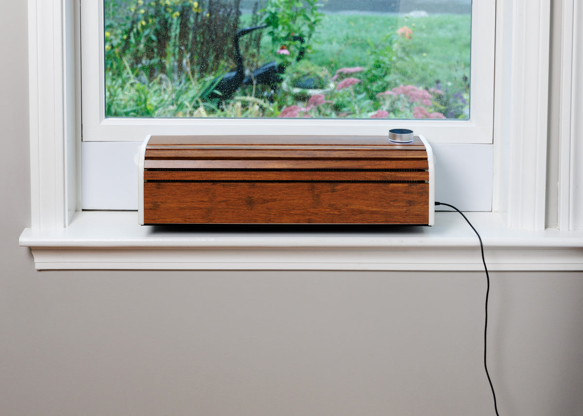 A rectangular wooden portable window air exchanger energy recovery ventilator ERV with a warm brown wood-grain finish and white side trim, resting on a white windowsill with a garden view outside.